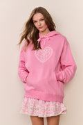 Woman wearing a pink hoodie with a heart design and a matching skirt against a beige background