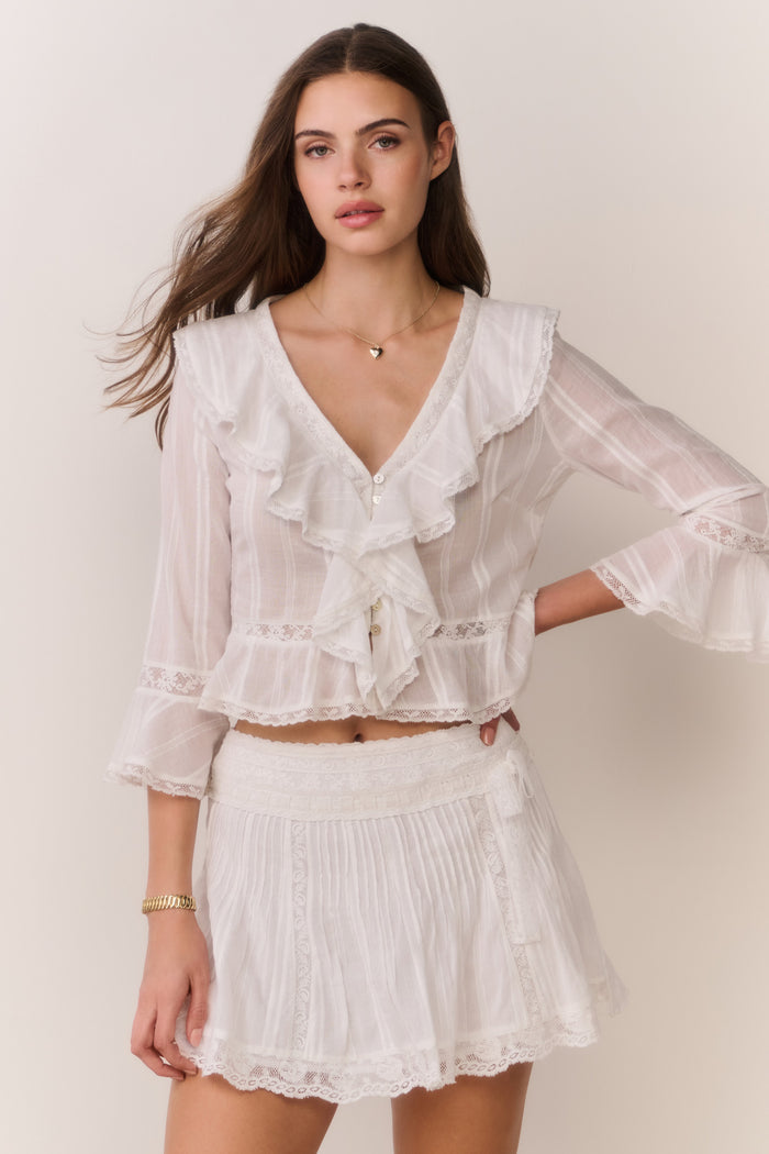 Woman wearing a white lace long sleeve ruffle trim blouse and skirt against a plain background