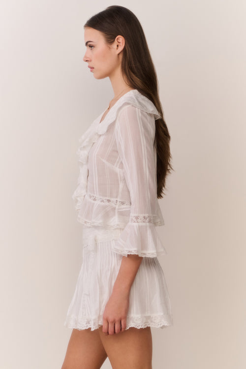 Woman wearing a white lace long sleeve blouse with ruffle trim and matching skirt against a plain background