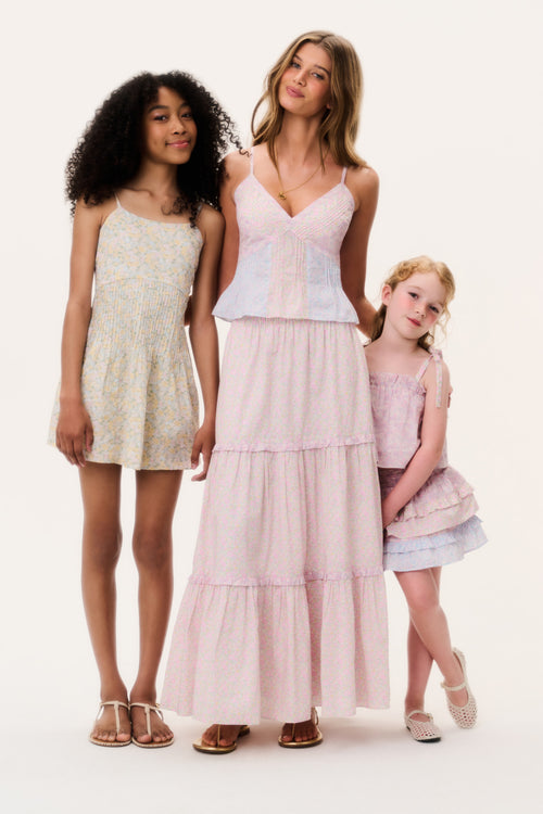 A woman and two young girls wearing light pink and floral dresses on a white background