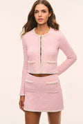 A woman modeling a light pink tweed two-piece set consisting of a cropped long-sleeve jacket with cream trim and a matching mini skirt, against a solid light beige background.