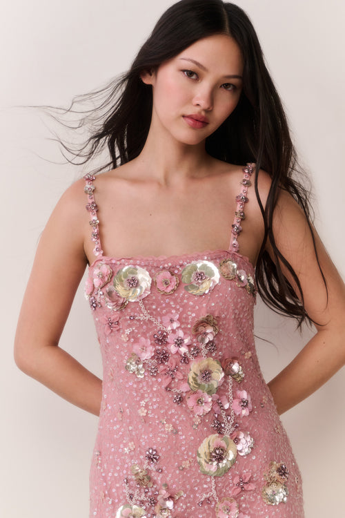 Woman wearing a pink dress with floral embellishments against a plain background