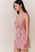 Woman wearing a pink dress with floral embellishments against a plain background