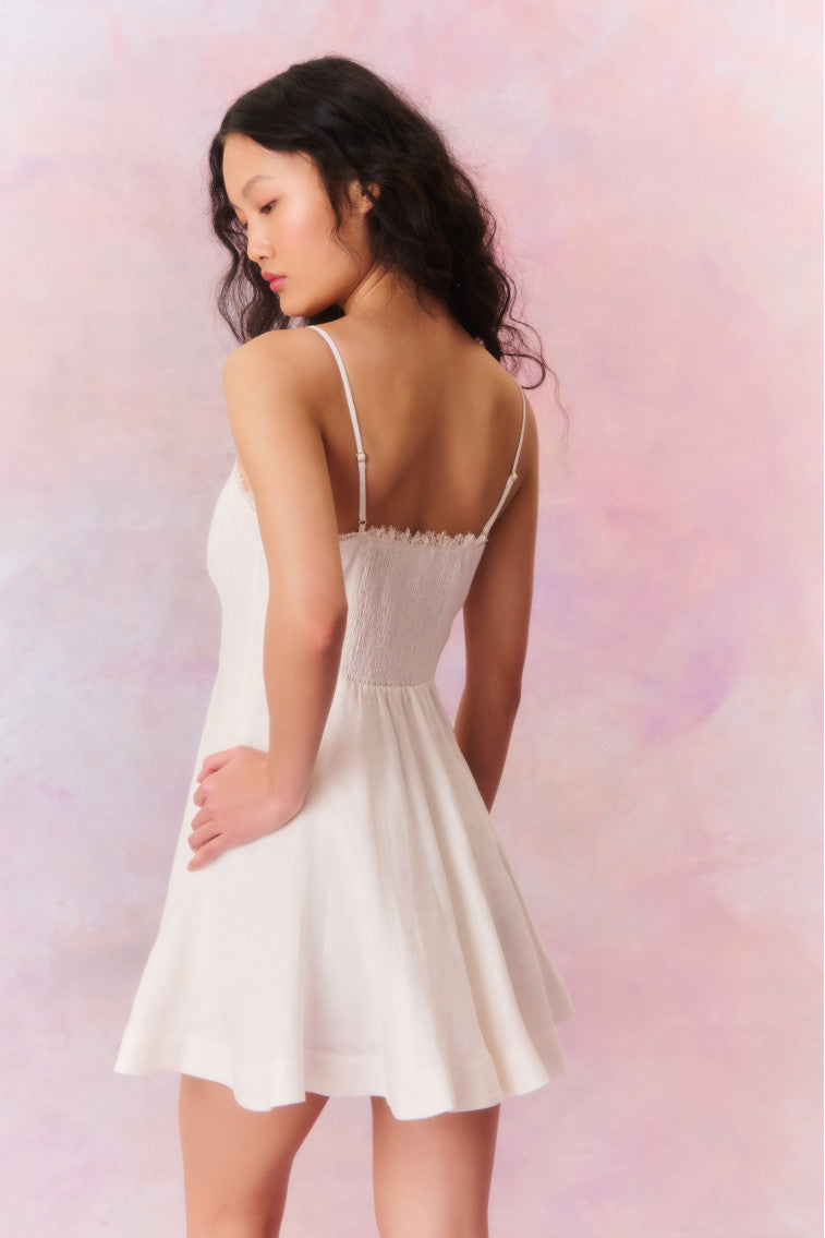 White mini dress with princess seams down the waistline that releases into a full sweepy skirt. Includes a smocked back, cups with shirring details, and delicate lace along the neck’s edge.