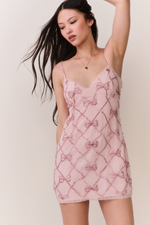 Woman wearing a pink lace mini dress with bow pattern embellishments on a white background
