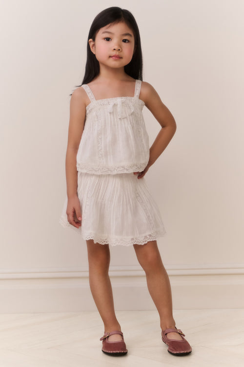 Little girl wearing a white lace trim camisole and matching skirt against a plain background