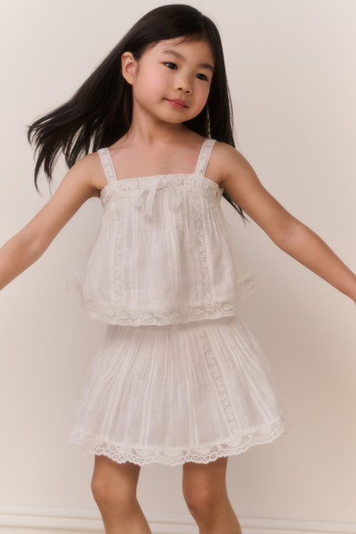 Little girl wearing a white lace trim camisole and matching skirt against a plain background
