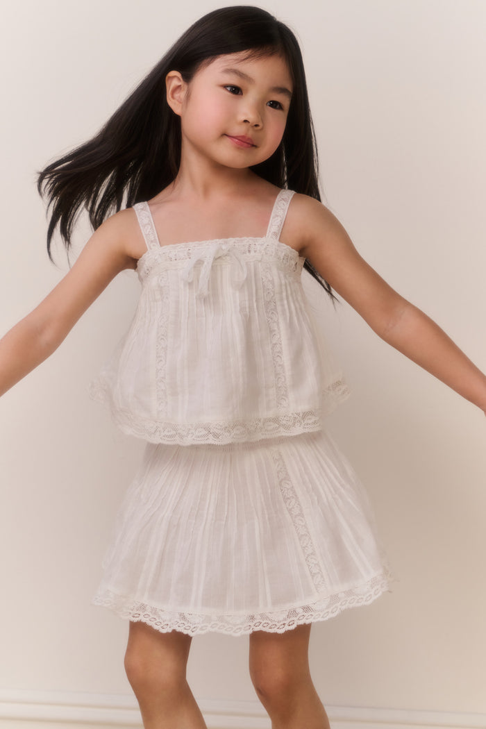Little girl wearing a white lace trim camisole and matching skirt against a plain background