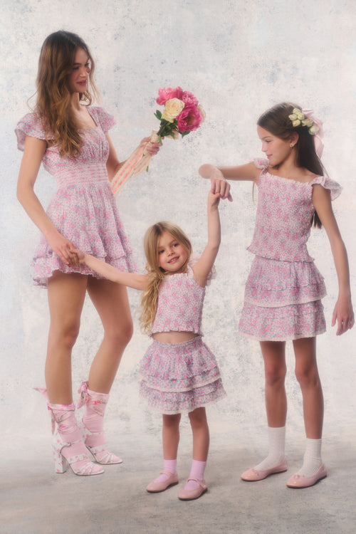 Three women in matching pink outfits with floral accessories on a light background