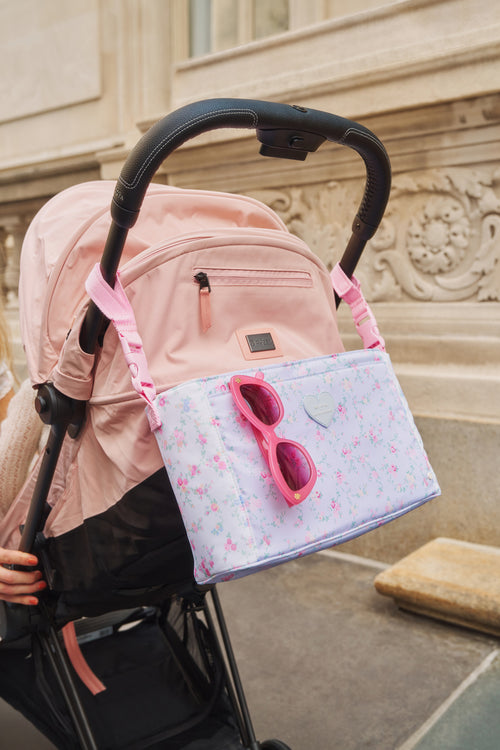 Pink backpack with sunglasses attached to a stroller outdoors.