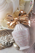 White LoveShackFancy heart perfume bottle with gold bow, pearls and floral decor.