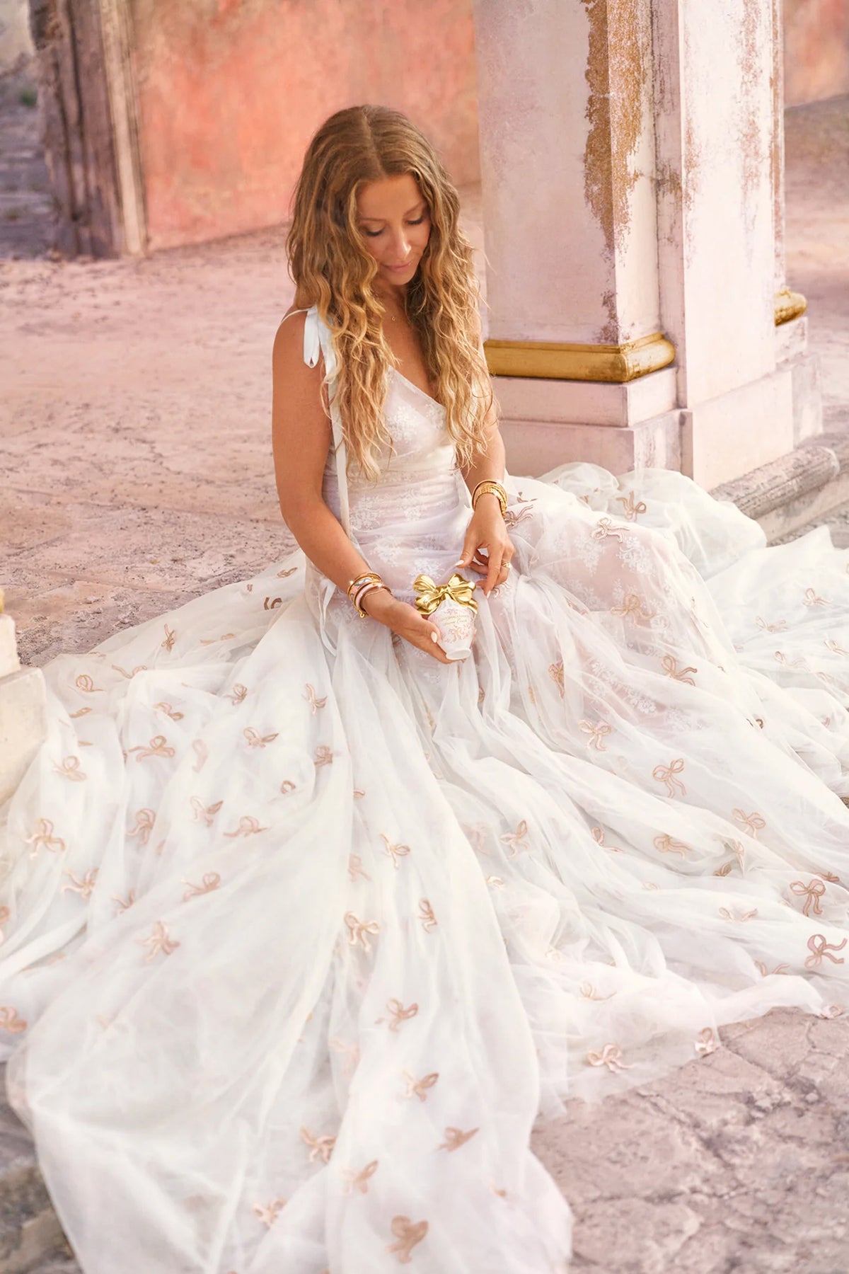 Model in white dress holding LoveShackFancy vanity bottle with gold bow topper.