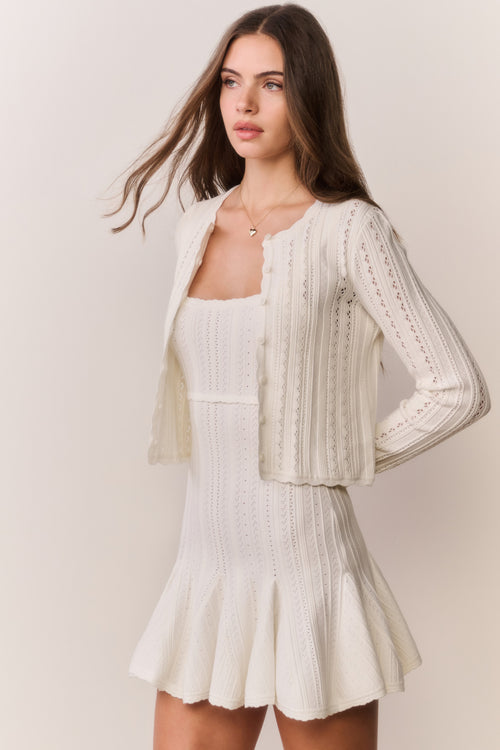 Woman wearing a cream-colored cardigan and dress with intricate patterns on a plain background