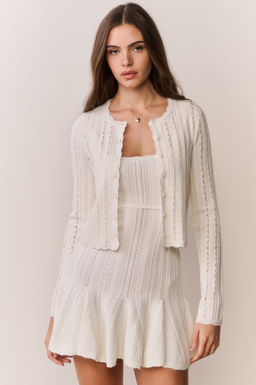 Woman wearing a cream-colored cardigan and dress with intricate patterns on a plain background