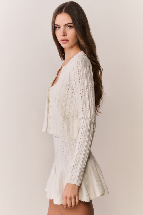 Woman wearing a cream-colored cardigan and dress with intricate patterns on a plain background