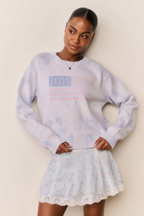 Woman wearing a light blue sweater with an American flag design and a floral skirt against a beige background