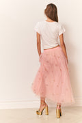 Person wearing a white t-shirt and pink tulle skirt with gold shoes on a plain background