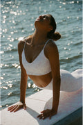 Woman in a white bikini sitting by a body of water with sunlight reflecting on the surface.