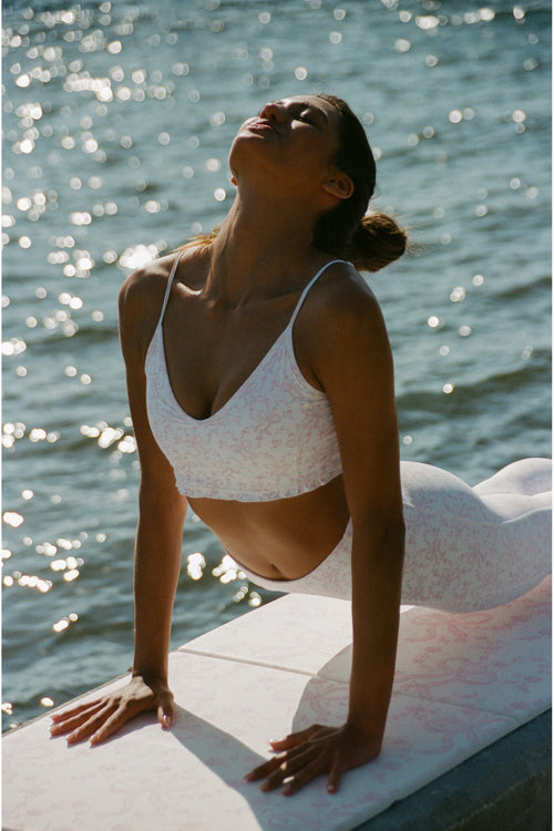 Woman in a white bikini sitting by a body of water with sunlight reflecting on the surface.