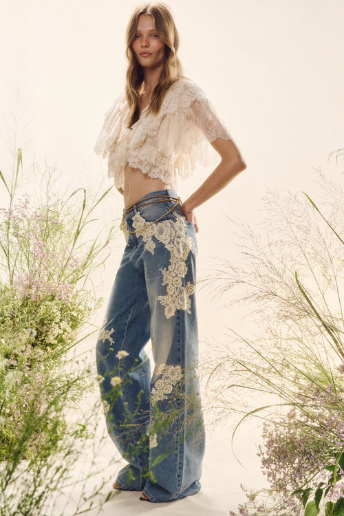 Woman wearing a white lace top and blue jeans with floral patterns, standing in a natural setting with flowers.