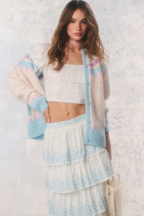 Woman wearing a pastel cardigan over a white top and skirt with a textured white background