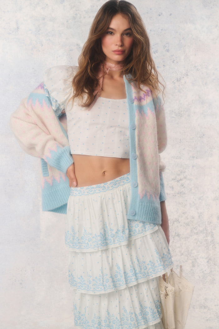 Woman wearing a pastel cardigan over a white top and skirt with a textured white background
