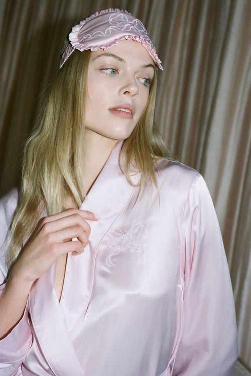 Woman wearing a pink satin robe and matching headband against a beige curtain background
