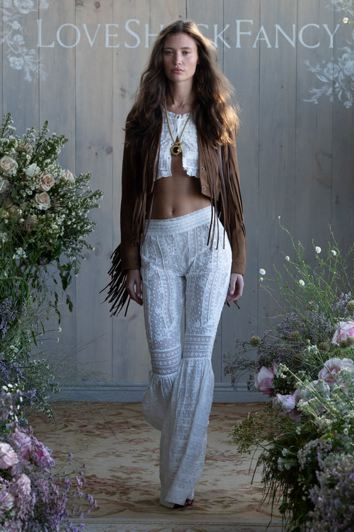 Woman in a Brown suede fringe jacket and lace white pants with floral decorations and 'LOVE SHACK FANCY' branding.