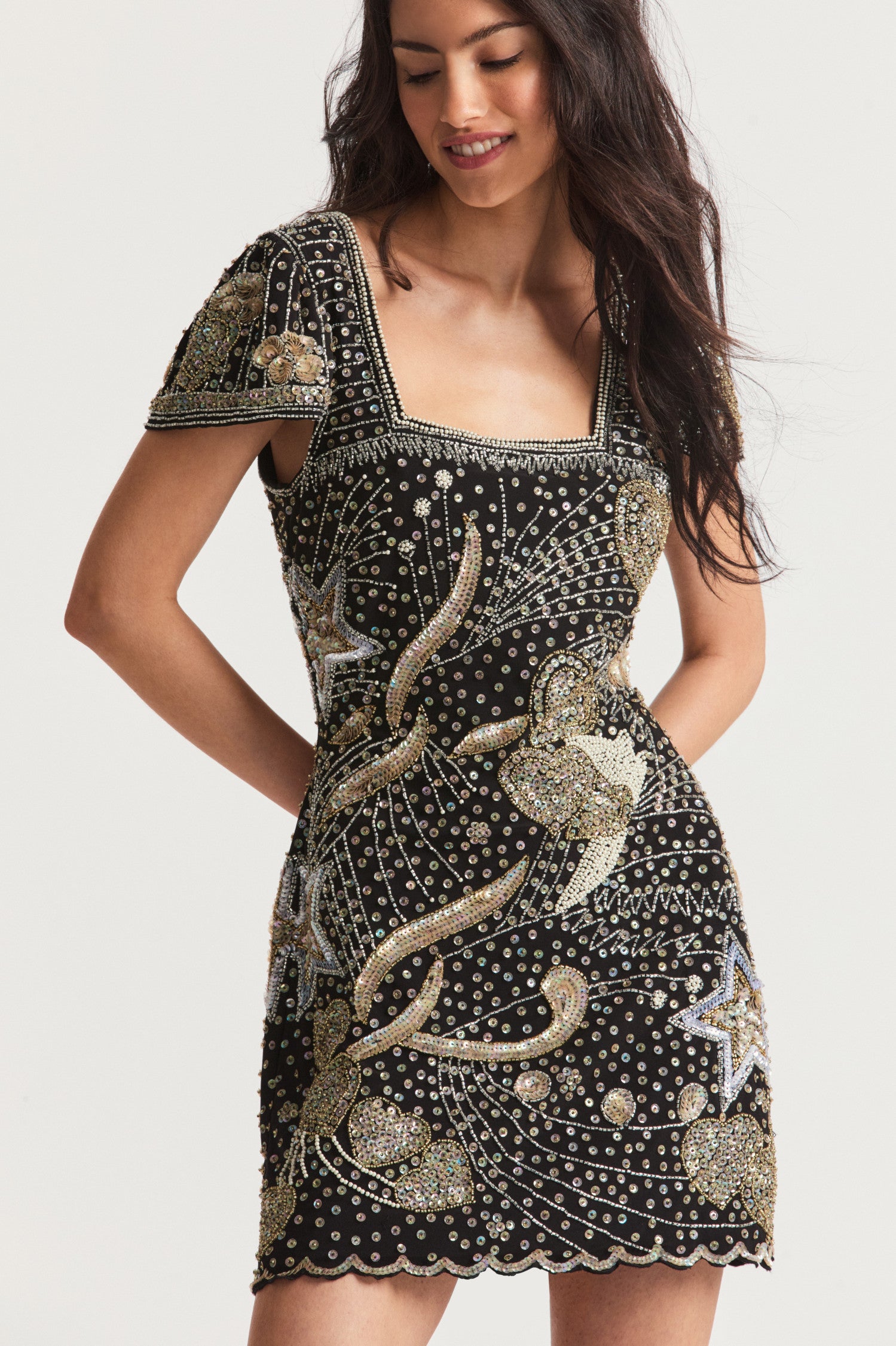 Black mini dress with iridescent gold and silver sequins outlining an arrangement of whimsical hearts and stars with wisps and sprays of shimmer all over on a stretchy crepe fabric for easy wear. Fully lined, this dress features a square neckline, cap sleeves, a fitted style, and a small sweep with a sequined scalloped finish.