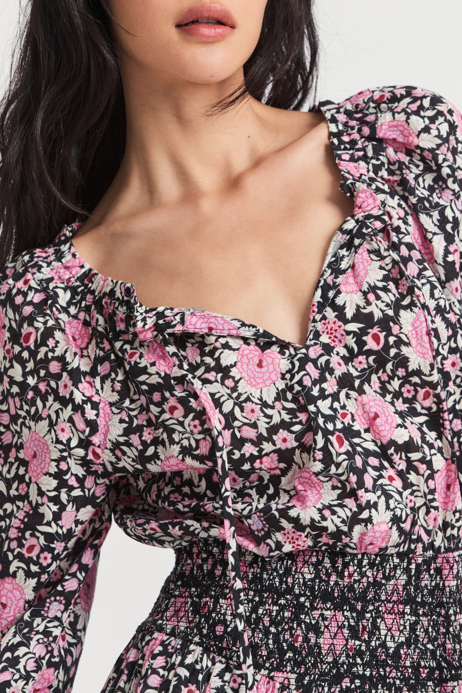 Black mini dress with a pink floral print. Featuring an elasticated neck opening with a tie at the front, an easy bodice with a stretchy smocked waist that releases into a tiered skirt that hikes up slightly on each side.