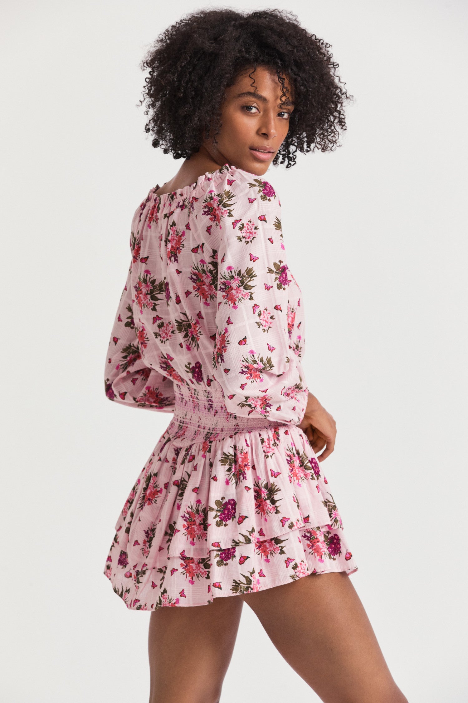 Pink mini dress with a pink floral print. Featuring an elasticated neck opening with a tie at the front, an easy bodice with a stretchy smocked waist that releases into a tiered skirt that hikes up slightly on each side.