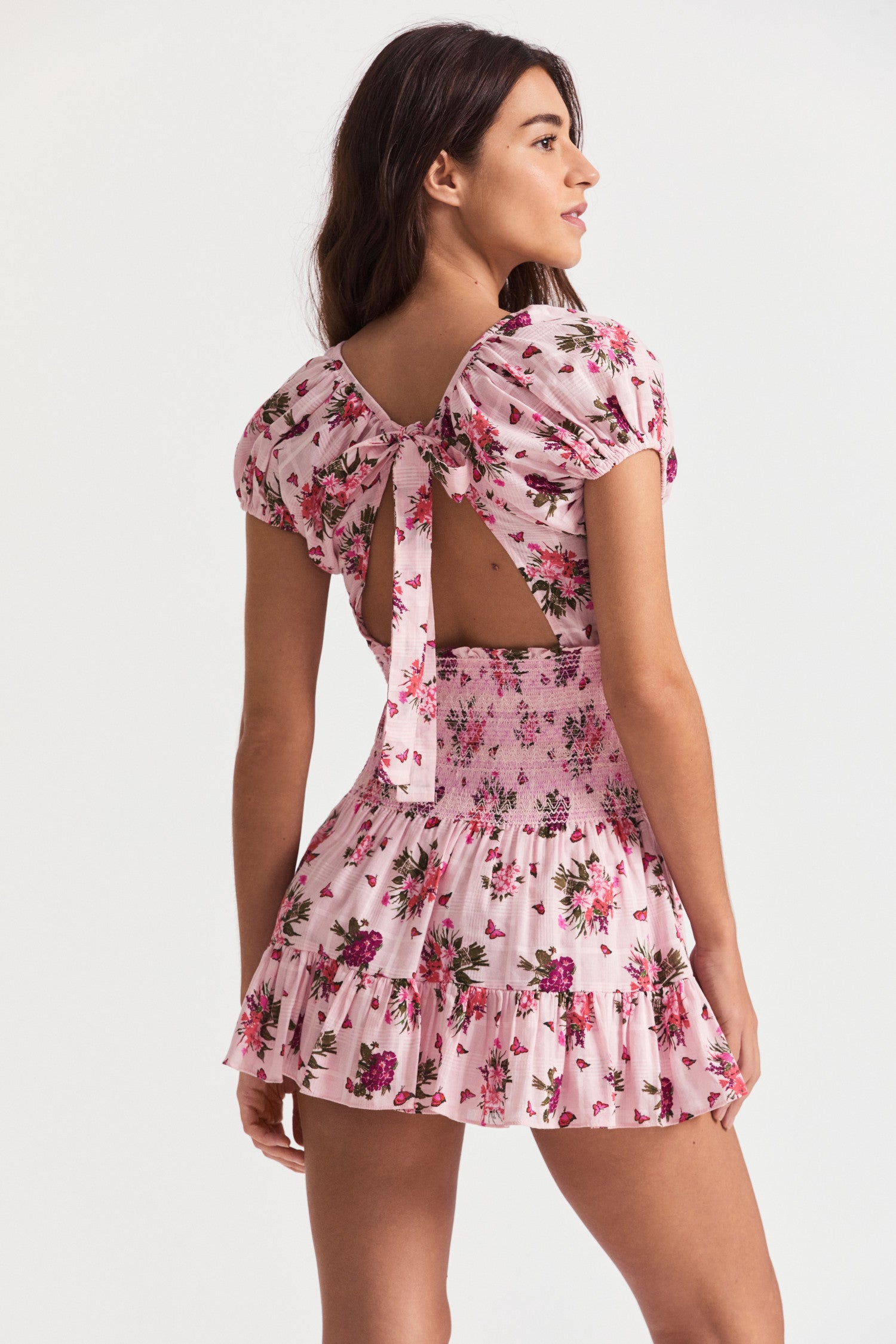 Pink floral mini dress with smocking detail, an elasticated neck opening, short puff sleeves, a flowy skirt, and a bow revealing a slightly open back. 